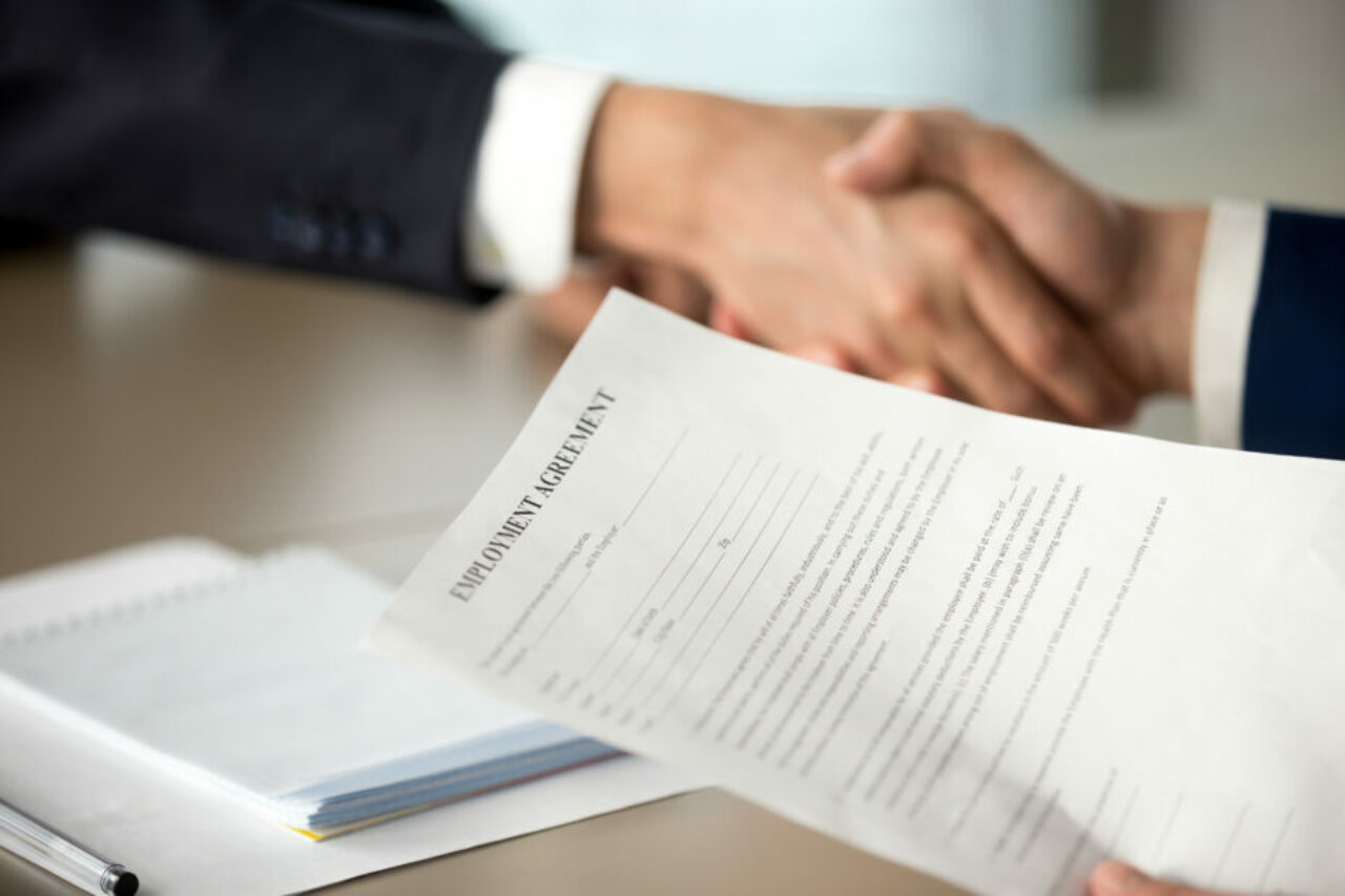 Close up photo of employment agreement document in hand of hiring manager shaking hand of job candidate. Employer offering work contract to applicant, successful interview, positive hiring decision